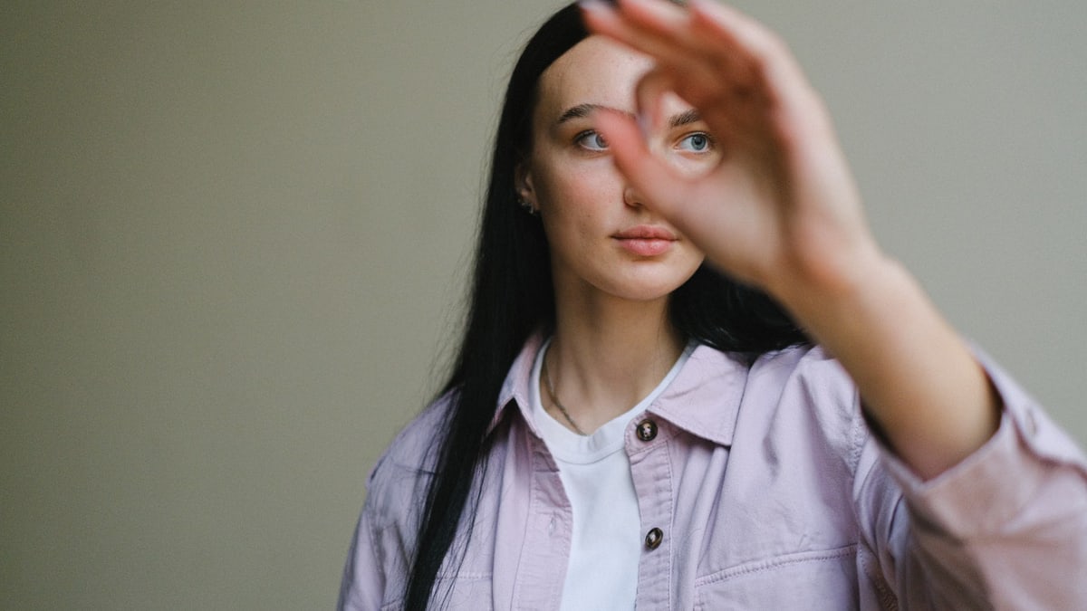 Content young female in trendy shirt looking away through ok gesture on gray background of studio